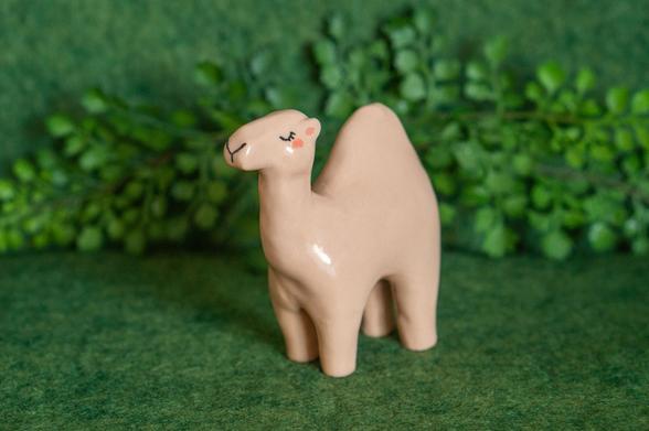 A minimalistic dromedary camel ceramic figurine. The camel is light brown and in a standing pose, with a peaceful expression with its eyes closed, featuring long eyelashes.