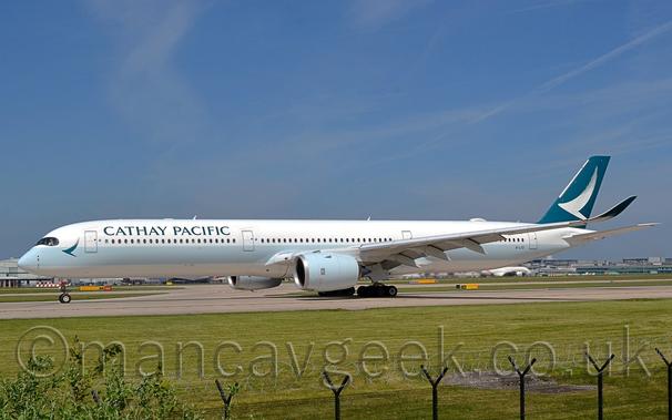 Side view of a white, twin engined jet airliner parked on a runway facing to the left.
The plane has a very pale blue, thick stripe running along the body under the passenger cabin windows, with a green stylised flying bird that looks as if drawn by a single stroke of a brush behind the cockpit, and green "Cathay Pacific" titles on the upper forward fuselage.
The tail is green with a larger white version of the bird logo.
The upturned wingtips are also green, this time with just the tips of the birds wings bleeding through from the white underside of the planes wings.
The engine pods slung under the wings are the same pale blue as the body stripe.
A wide strip of green grass dominates the foreground, with a black mesh metal fence stretching across the bottom of the frame.
In the distance, grey hangars can be seen under the planes nose, with a wide grey terminal under the mid and rear fuselage,
Blue sky with smears of thin grey cloud fill the rest of the frame.