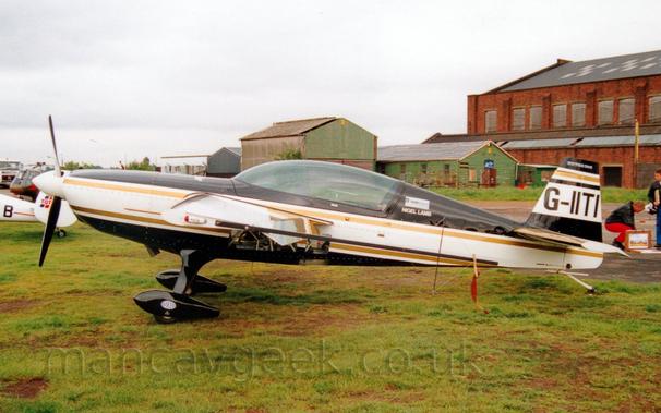 Side view of a white and black, single engined aerobatic light aircraft parked on grass facing to the left.
The plane has black top and bottom surfaces, with a thick white stripe running along the body, and gold piping in between.
The registration "G-IITI" is on a thick white stripe on the black tail.
There is a large, glazed canopy over the cockpit, with small white "Nigel Lamb" titles on the fuselage immediately behind the canopy.
There are also glazed panels in the lower fuselage, under the wings.
Black spats cover the wheels on the fixed landing gear.
Green grass fills the foreground.
A large, red brick hangar is on the right of the frame in the background, with smaller green buildings next to it, towards the middle of the frame.
The tail of a white light aircraft and a small, camouflaged helicopter are in the background on the left.
Cloudy grey sky fills the rest of the frame.