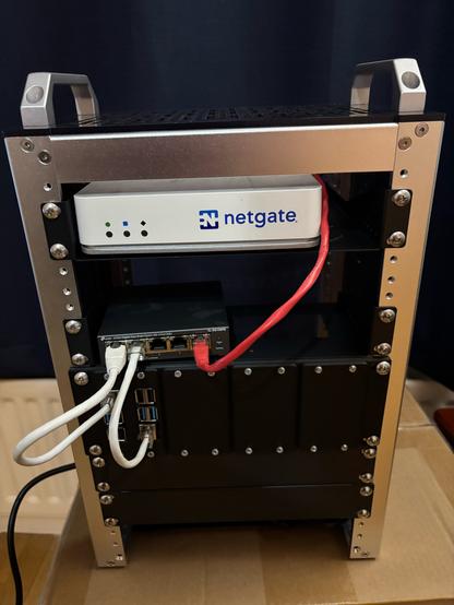 A picture of an 8U mini rack. One view is taken up with a Netgate firewall. A space is left to install a keystone patch panel. When you is taken up by one 5 port switch. 2U is taken up by a panel that can accommodate up to 8 Raspberry Pis, only two are currently installed. Blanking panels make up the next 2U of space, with 1U missing due to not having enough screws. A red cable can be seen coming out of the firewall and into the switch this is temporary until the Keystone panel is installed. Two more ethernet cables can be seen coming out of the switch into the raspberry pis