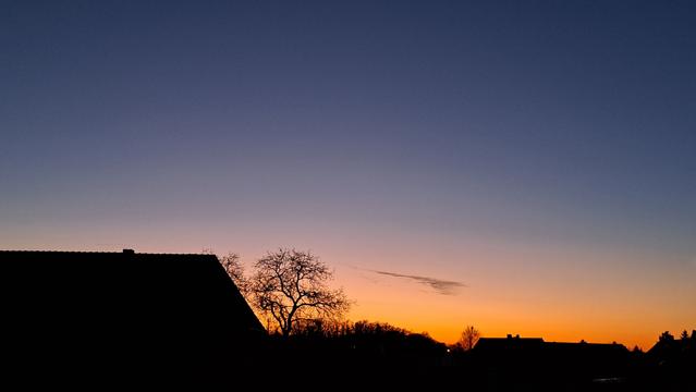 Fensterblick: Dunkelblauer Abendhimmel mit einer kleinen grauen Schleierwolke überm Horizont, welcher leuchtend orangerot eingefärbt ist. Silhouetten von Hausdächern und Bäumen am unteren Bildrand.