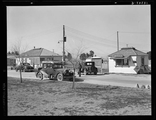 The image depicts a black and white scene from what appears to be the early 20th century, likely captured by Dorothea Lange for her work with the Farm Security Administration (FSA). The setting is an outdoor area near a rural road where several vintage automobiles are parked or driving. In the foreground, there's a person standing beside one of these cars; their gender and race aren't distinguishable from this distance.

The middle ground shows buildings that seem to be part of a camp for migratory laborers at Indio in California, with signage such as "OFFICE" indicating some administrative structures or services. The architecture is modest, typical of mid-20th-century farm settlements.

Behind the people and vehicles stands an open field with sparse vegetation under a clear sky, suggesting arid conditions consistent with Coachella Valley's climate at that time. Telephone poles line the road, which implies connectivity to other parts of California or beyond during this period when such infrastructure was less widespread in rural areas.

The overall mood is quiet yet contemplative, capturing daily life and work amidst agricultural labor settings as documented by Lange through her lens.