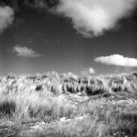 Black and white photo 6x6 of a dune on the beach of the Baltic Sea. The blue sky is rendered very dark, individual white clouds. The dune is grassy.