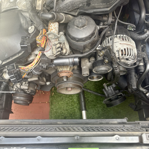 The front of the M54B25 inline six-cylinder engine in my 2003 BMW 525iT wagon, with the cooling system and accessory belts removed. The air conditioning compressor, on the left, and power steering pump, on the right, both hang suspended by zip ties from the inner fenders, and the plugs of the engine wiring harnesses lay loose atop the engine in preparation for removal and replacement of the engine and transmission.