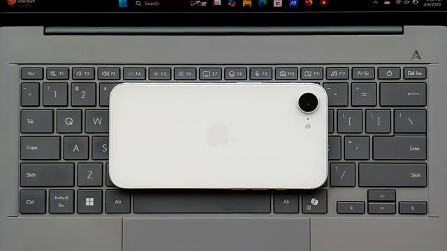 Photo of a white Apple iPhone 16e, face down on the keyboard deck of a gray ASUS Zenbook A14 laptop.