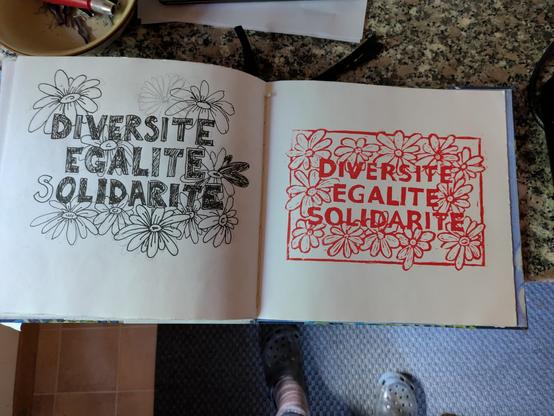 Ein offenes Skizzenbuch. Auf der linken Seite mit filzer skizziert: DIVERSITE, EGALITE, SOLIDARITE. Rundherum Blümchen. Auf der rechten Seite der fast fertige Linoldruck in rot, der gleiche Schriftzug, wieder mit Blumen rundherum. Einige feinheiten an der Druckplatte möchte ich noch etwas korrigieren.