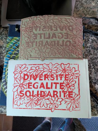 Der Linolschnitt und darunter ein fertiger Druck in rot. DIVERSITE, EGALITE, SOLIDARITE umschlossen von Margeritenähnlichen Blumen.