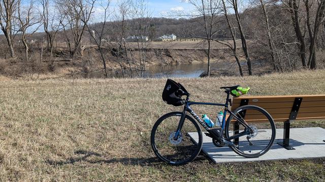 Blue and black Roubaix bicycle leaning on a park bench off the Zumbro South Trail looking Southeast with the River behind and in the distance the Mayowood Stone Barn complex with hills behind under mostly blue skies.