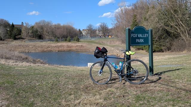 Bleu and black Roubaix bicycle leaning on the green "Fox Valley Park" sign with a  retention pond behind and the park with a ball field in the distance along with neighborhood homes under blue skies.