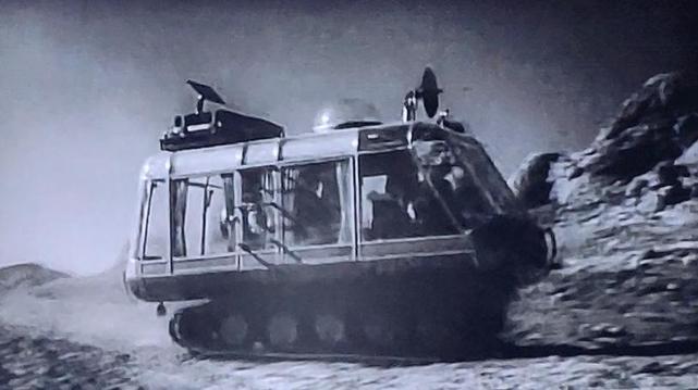 Their vehicle is sort of like a large glass van with lots of seats. Instead of wheels it has the rollers of a tank. Shown in black & white film. The vehicle is on a rocky terrain.