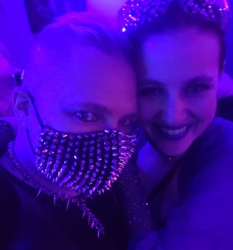 Two people's faces. One in a spiked face mask both in goth makeup,  under a purple light