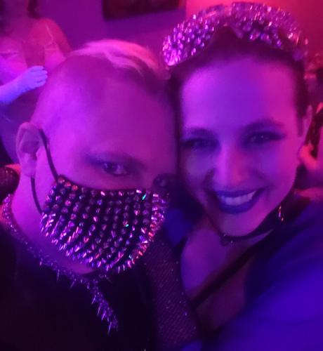 Two people's faces. One in a spiked face mask both in goth makeup,  under a magenta light