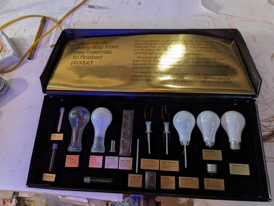 A display box from GE on how they maka a lightbulb. Various glass stems and bulb shapes are in the box going from left to right in various steps of completion with a gold foil descibing how GE controls every step of the process.