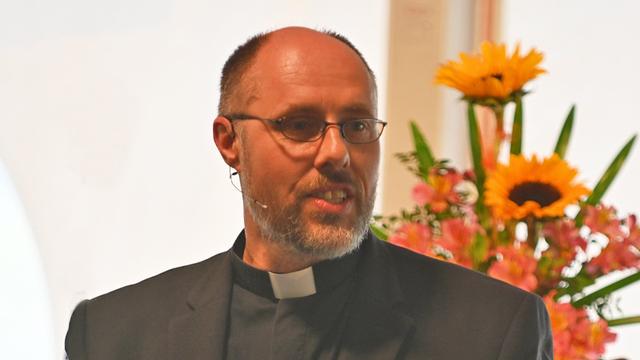 Porträt von Hendrik Walz. Ein Mann mittleren Alters mit Brille und schwarzem Sakko. Er trägt ein schwarzes, geöffnetes Kirchenhemd und spricht. Im Hintergrund sind Sonnenblumen in einem Blumenstrauß zu sehen. Der Mann wirkt konzentriert und engagiert.