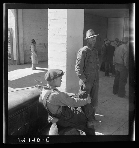 The image depicts a group of men in outdoor industrial clothing, likely farm laborers or workers from the early to mid-1900s. Two prominent figures are sitting on what appears to be an agricultural conveyer belt system commonly used for sorting crops such as peas during harvest time; this is suggested by their overalls and caps indicative of rural work attire.
The men's expressions range from contemplative to weary, reflecting perhaps a momentary pause in their labor or the fatigue associated with manual fieldwork. In the background stands another figure wearing lighter-colored clothing and headgear, possibly overseeing the operation or simply passing through this communal space.
A large concrete structure dominates one side of the photo, suggesting an industrial setting such as part of a processing plant near agricultural fields. The black-and-white quality of the photograph adds to its historical context, likely taken during World War II given the caption reference and era-specific attire worn by individuals in it.

The image captures a moment that may be indicative of social issues related to labor rights or poverty prevalent at the time; such depictions are often found within works associated with Dorothea Lange.