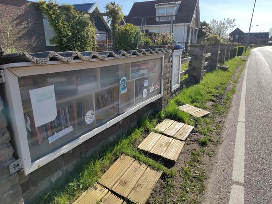 Minibieb en weggeefkastje naast elkaar langs een berm langs een smalle straat. Op het gras in de berm liggen voor de biebje en het kastje houten vlonders