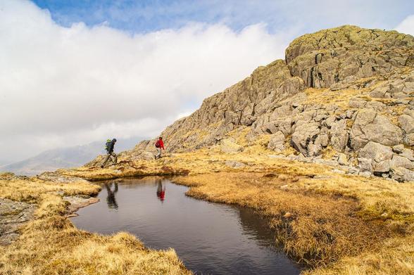 Small figures of two walkers/climbers are mirrored in a small mountain tarn. They’re heading towards rocks which rise quite steeply on the right side of the picture.