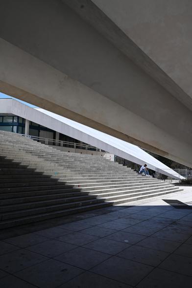 A modern architectural space dating back from the 60`s in the GDR/ Berlin Alexanderplatz/ at the entry level of the tower, with wide concrete steps and geometric overhangs casting shadows. A person is sitting alone on the steps near the right side of the image, partially lit by sunlight. The scene conveys a mix of light and shadow, emphasizing the structure’s lines and angles.