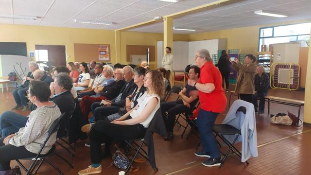 Des personnes assises en trois rangées assistent à une réunion publique dans une salle d'école.