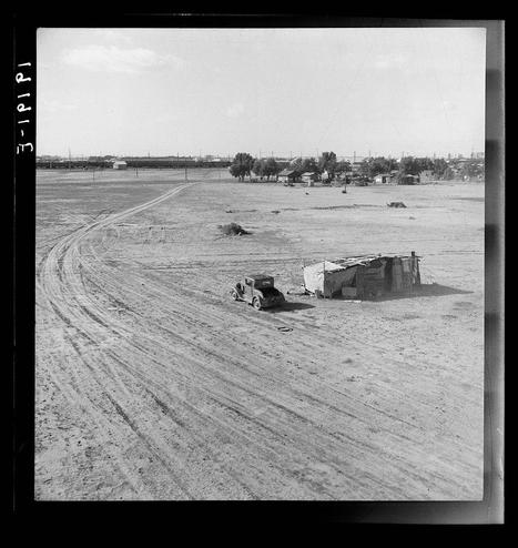 The image depicts a rural landscape with tracks in the dirt leading towards several makeshift structures and vehicles. The scene appears to be from an agricultural area, possibly during or after harvest season given the presence of tractors and trailers used for farming purposes.
There are multiple small buildings made up of various materials such as metal sheets, wood planks, and tarpaulins that suggest temporary living arrangements typically found in migrant labor camps. In the background, there is a more permanent structure with a pitched roof which might be part of a larger farmstead or community facility like an FSA migratory labor camp.
The black-and-white photograph captures a stark contrast between the arid landscape and the simple sheltered dwellings. The overall composition conveys a sense of hardship faced by migrant workers, as well as their resilience in adapting to conditions with limited resources.
This image is associated with Dorothea Lange's work during her time documenting American life through photography for the Farm Security Administration (FSA), which was established to support and assist farmers facing financial difficulties. This particular photograph falls within a series that captures moments of Mexican families living as migratory workers in California, particularly highlighting their challenges due to displacement or seasonal labor demands.