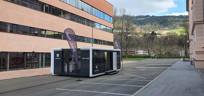 Das Foto zeigt den Ausstellungspavillon des Technikunternehmens Zwick-Roell auf dem Gelände der Montanuniversität Leoben in Österreich. Der in weiß und schwarz gehaltene mobile Container kann auseinandergefahren werden und bietet 40 Quadratmetern Ausstellungsfläche. Vor und hinter dem Container stehen zwei Flaggen mit dem Firmennamen Zwick-Roell