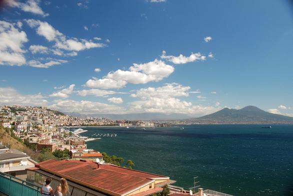 Photo showing view across the Gulf of Naples from the edge of the Phlegraean Fields caldera crater to Mount Vesuvius. The sky is blue and there is dark emerald-green water between settlements with terracotta roofs and a dark double-peaked mountain on the horizon. Photo thanks to Gerhard Wörner