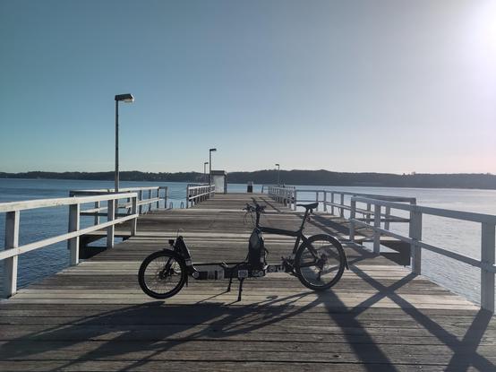 Ein Bullitt Lastenrad, welches quer auf dem Anleger Bellevue in Kiel steht. Im Hintergrund die Förde. Morgendliche Sonne auf der rechten Bilddecke