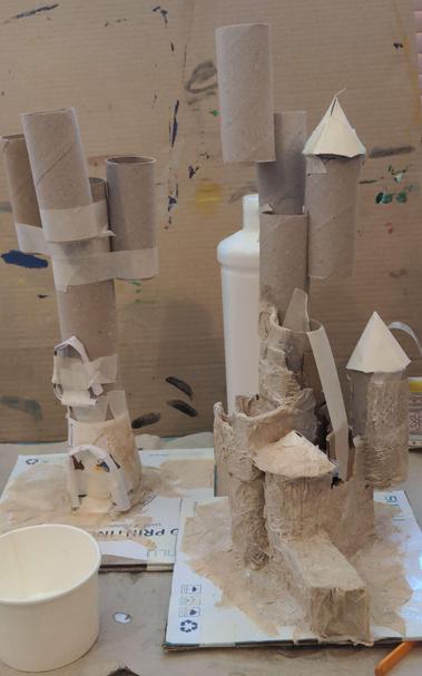An in progress shot of two magical castles being built from card and loo roll, on a messy desk.