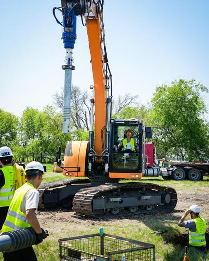 Two team members in yellow safety vests are carrying parts of the tunneling machine to the center where an orange crane is lifting another part of the machine.