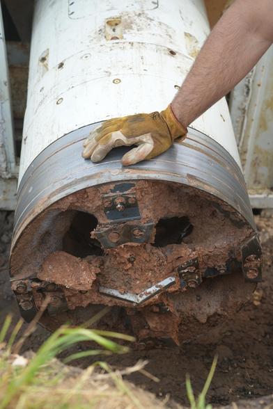 A close-up of a tunnel boring machine drill head covered in red-brown soil, with a gloved hand resting on the metal casing.