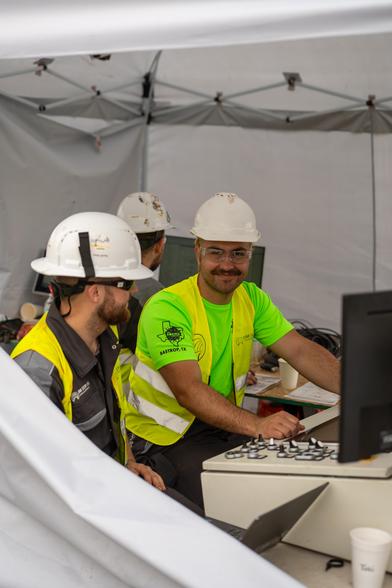Two team members of TUM Boring wearing safety gear and helmets working at a control station under a tent, focused on the screens in front of them.