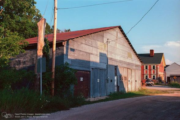 A film photograph taken at ground level depicts a rural scene at dusk, featuring a large, weathered barn as the primary subject. The barn is constructed of corrugated metal siding with a red roof and is positioned on the left side of the frame. The setting sun casts long shadows across the barn's surface, highlighting its texture and age. To the left of the barn, lush green foliage and tall grass grow along the edge of a gravel road. A utility pole stands near the barn, with wires stretching across the clear blue sky. In the background, a red wooden house with weatherd paint and white-framed windows and a black roof is visible, adding depth to the composition. A silver SUV is parked in front of the house. The overall mood of the photograph is tranquil and nostalgic, evoking a sense of rural Americana. The image captures the beauty of the aging structures and the natural landscape, emphasizing the quiet charm of the countryside.
