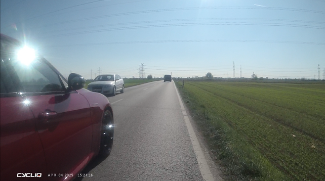 Eine Landstraße, rechts davon ein grünes Feld, blauer Himmel.
Sehr nah am Fahrbahnrand die Beifahrerseite eines Autos.
In der Mitte der Straße gestrichelte Linien.
Auf der Gegenfahrbahn ebenfalls ein Auto.