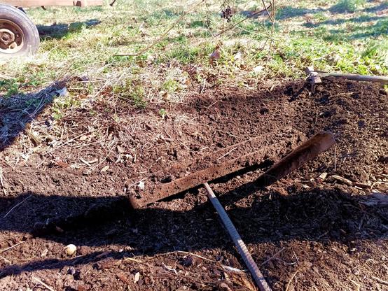 A prybar, lifting up a steel I-beam that is still partially buried and covered in soil. Freshly dug soil is spread around and an old rake lies on the ground behind. Part of a tractor trailer tyre is visible further into the grass.