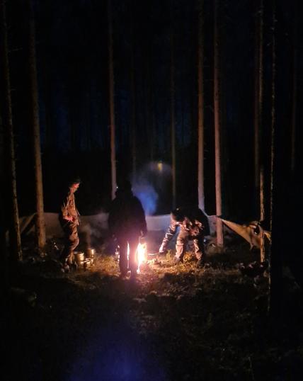 Kolme ihmistä nuotion ympärillä pimeässä metsässä. Puihin heidän ympärilleen on viritetty sadeviitat laavuiksi.