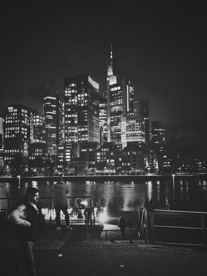 The image is a black and white portrait-orientated photograph taken at night. It shows a waterfront scene with a cityscape in the background.
In the foreground, there is a paved walkway with a metal railing along the water's edge. Several people are visible on the walkway. One person is entering the frame and walking towards the right side of the frame. Behind that person another one is facing away from the camera, looking towards the cityscape and the river. 
Behind the walkway and the water, there is a dense cluster of tall buildings illuminated with numerous lights from their windows. The buildings vary in height and architectural style. The lights reflect on the surface of the water, creating streaks of brightness. The sky above the city is dark and overcast.