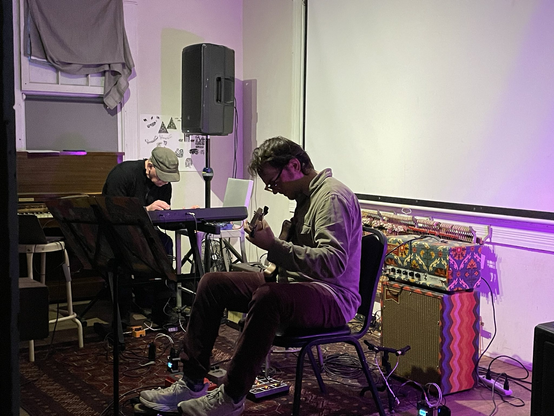 Wayne Horvitz, an older white man dressed in a dark sweater and brown cap, sits raptly at his keyboard in the background to the left of the center of the picture. In the foreground, Anthony Pirog, a younger white man with dark hair and glasses, wearing a grey button-down shirt, plays guitar at the center of the picture in the foreground. The are playing in a small room. There is a blank movie screen drawn on the wall behind them, with speakers, amplifiers, and a piano also in the room.