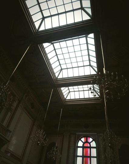 Eine Prunkaula, mit Marmor(?) -verkleidung, drei Oberlichter in einer schicken Holzkasettendecke. An der Stirnseite ist ein buntes Glasfenster. Kronleuchters. Ein erhellender Filter auf dem Bild.
