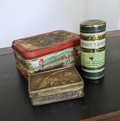 Trois boîtes posées sur le cuir vert d'un bureau. Une grosse boîte rectangulaire un peu cabossée, rouge, décorée des dessins des fables de La Fontaine. Une petite boîte au couvercle de laiton embossé de feuilles et fruits de platane (?). Une haute boîte cylindre au décor napoléonien vert et or de charbon naphtolé "puissant désinfectant de l'estomac et de l'intestin".