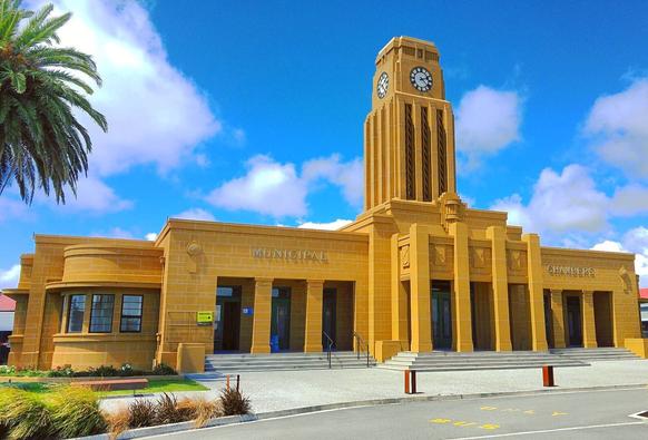 Photo shows a large yellow two wing building with a tall clock tower in the centre. It is a warm day with a lightly cloudy deep blue sky. In front of the building there are a concrete area with some small. The change to the sidewalk are marked with colourful small poles. To the left there are a green turf with some garden furniture, and a palm tree frames to the left side of the photo.