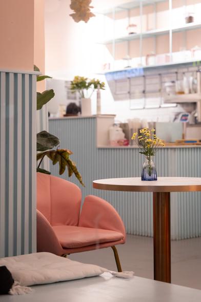 A cozy pastel-colored café interior with a soft pink armchair, a round table holding a small vase of yellow flowers, and a light blue striped counter in the background. The setting is softly lit, creating a calm and inviting atmosphere.