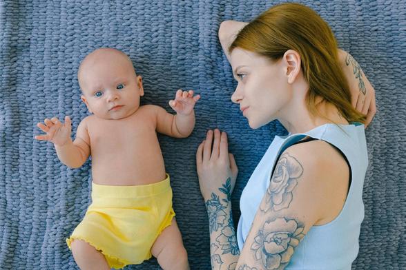 Ein Baby mit blauen Augen und hellgelbem "Windelröckchen" und eine an den Armen mit Blumenmustern tätowierte junge Frau mit rötlichem Haar und hellblauem, ärmellosem Oberteil liegen auf einer blauen Decke. Die Mutter ist dem Kind zugewandt.