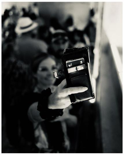 This black and white image depicts a woman holding a smartphone, capturing a selfie or video in a public setting with other people in the background. Her smartphone case has slots with credit cards visible, focusing attention on personal details typically stored within phone cases. The background is slightly blurred, emphasizing the action of the woman and her smartphone. The composition conveys a candid, social moment, likely at a gathering or event.