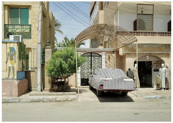 The image depicts a street scene that appears to be in a Middle Eastern setting, characterized by its architecture and elements. Visible in the scene are:

1. **Buildings**: There are multi-storied buildings with features typical of Middle Eastern architecture, such as ornate window grilles and balconies.

2. **People**: Two fashion dummies can be seen standing near the entrance of a shop. They are dressed in traditional attire, suggesting a cultural context.  

3. **Shop Signage**: The shop has Arabic text on its signage, indicating that the location could be in an Arabic-speaking country. The script on the shop can be translated or transcribed but I’m unable to provide specific translations unless the text is better resolved.

4. **Car**: There is a car parked on the street, covered with a tarp, indicating it might not be in use currently or is being protected from the elements.

5. **Decorative Elements**: An interesting decorative feature—an arched framework (possibly made of bamboo or a similar material)—is seen above the sidewalk, adding a unique aesthetic to the streetscape.

6. **Vegetation**: Small trees and plants are present, suggesting that this is a residential area or close to one.

Overall, the image captures everyday life in a quiet street, possibly in a residential or semi-commercial part of a town or city in the Middle East.
