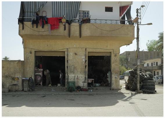 The image depicts a street scene with a two-story building. The ground floor has two open workshops or garages. Both appear to be cluttered with various items: one looks to have machinery or engines, and there is a presence of at least two individuals working or standing within. The other has numerous tires stacked both inside and outside, suggesting it might be a tire repair shop or similar business.

Above the shops, there is a residential floor with clothes hanging out to dry on a balcony guarded by wrought-iron railings. The building itself looks somewhat worn and has a simple architectural style. The area around the building is fairly barren, with minimal vegetation and more visible debris and scattered items on the street.

Additionally, utility poles and street infrastructure are visible, along with a partially visible building in the background, contributing to a scene that suggests a working-class neighborhood possibly within a busy, less affluent urban area.
