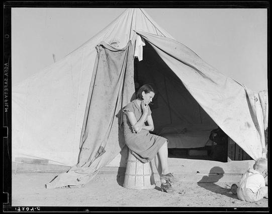 The image is a black and white photograph showing an outdoor scene with two subjects. In the foreground, there's a woman seated on what appears to be a wooden crate or stool in front of a large canvas tent that has some writing on it but is not fully legible due to the angle and distance.

She seems contemplative or possibly distressed, as she holds her arm near her face with her hand partially covering her mouth. She wears a dress from mid-thigh down to above the knees, and closed-toe shoes which are indicative of practical footwear suited for laborious activities.

Beside her on the ground is a young child sitting in what looks like an open cardboard box or similar container. The child's posture suggests they may be resting or simply observing their surroundings. Both subjects appear weary but there’s no direct interaction between them, giving off a sense of solitude despite being close to each other.

The setting seems arid and dusty with sparse vegetation indicative of desert conditions under bright sunlight. No distinct geographical features are visible apart from the sky which is clear without clouds indicating it might be midday or early afternoon due to the harshness of shadows cast on the ground by both subjects, suggesting a high sun position typical for regions like California.

Overall, this photograph captures an intimate moment in what appears to be part of a larger narrative involving migration and hardshi [...]