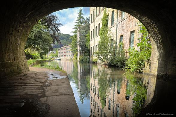 The image shows a picturesque scene of a canal viewed from under a stone bridge. The canal is flanked by old stone buildings with green windows, and there are trees and greenery along the water's edge. The water in the canal is calm, reflecting the buildings and trees.