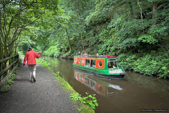 The image shows a person wearing a red jacket walking along a narrow path beside a canal. The canal is bordered by lush green trees and vegetation. On the canal, there is a colorful narrowboat painted in green and red, with the name 'Saracen' visible on the front. The boat has several people inside, and there is a bicycle on its roof.