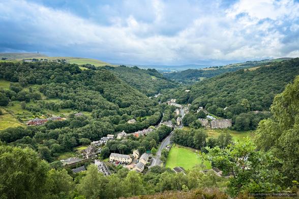 The image shows a picturesque valley with lush green hills and a small village nestled within it. The village has several buildings, including houses and what appears to be a few larger structures, possibly apartments or commercial buildings. A winding road runs through the village, connecting the various buildings and leading out of the valley. The hills surrounding the village are densely forested, and the landscape extends into the distance with more rolling hills and patches of open land. The sky is partly cloudy, adding to the serene and tranquil atmosphere of the scene.