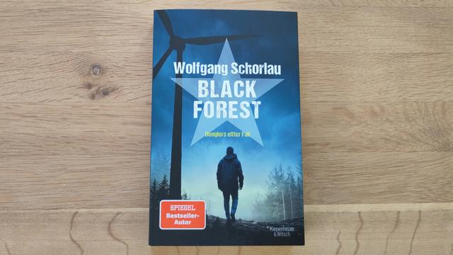 Das Buch liegt auf einer hellen Holzfläche. Das Cover ist überwiegend in Blau gehalten und ziert im unteren Bereich eine dunkle Fotografie einer hügeligen Waldlandschaft. In der Mitte des Vordergrunds sieht man die Rückansicht eines Mannes in einer Kapuzenjacke, der in Richtung Wald läuft. Links steht ein Windrad, das komplett schwarz ist, und rechts sind weitere Bäume des Waldes nur vage zu erkennen. Der mittlere Bereich des Bildes ist mit einem weißlichen Overlay versehen, und im oberen Bereich befindet sich ein transparenter, fünfzackiger Stern, auf dem in weißer Schrift der Autorenname steht. Darunter folgt der Titel, zweizeilig und in Großbuchstaben. Etwas weiter unten ist der Titelzusatz in gelblicher Schrift zu sehen. Unten links befindet sich der SPIEGEL-Bestseller-Aufkleber, und am rechten Rand steht der Verlagsname.